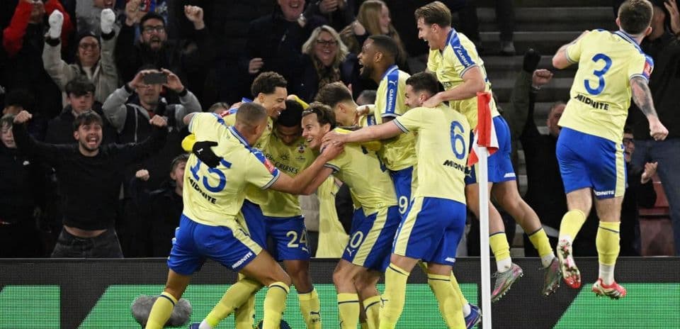 Jugadores celebrando un gol con aficionados de fondo