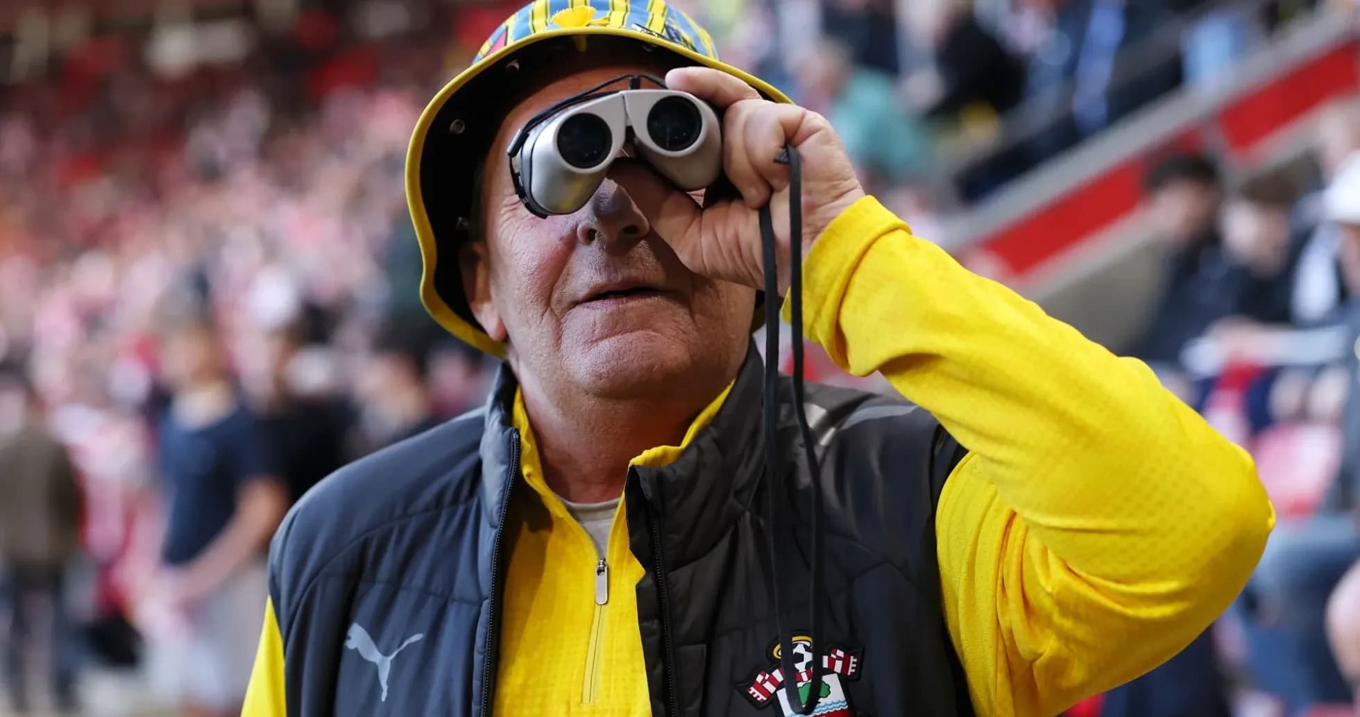 A fan wearing binoculars and a hat at stadium
