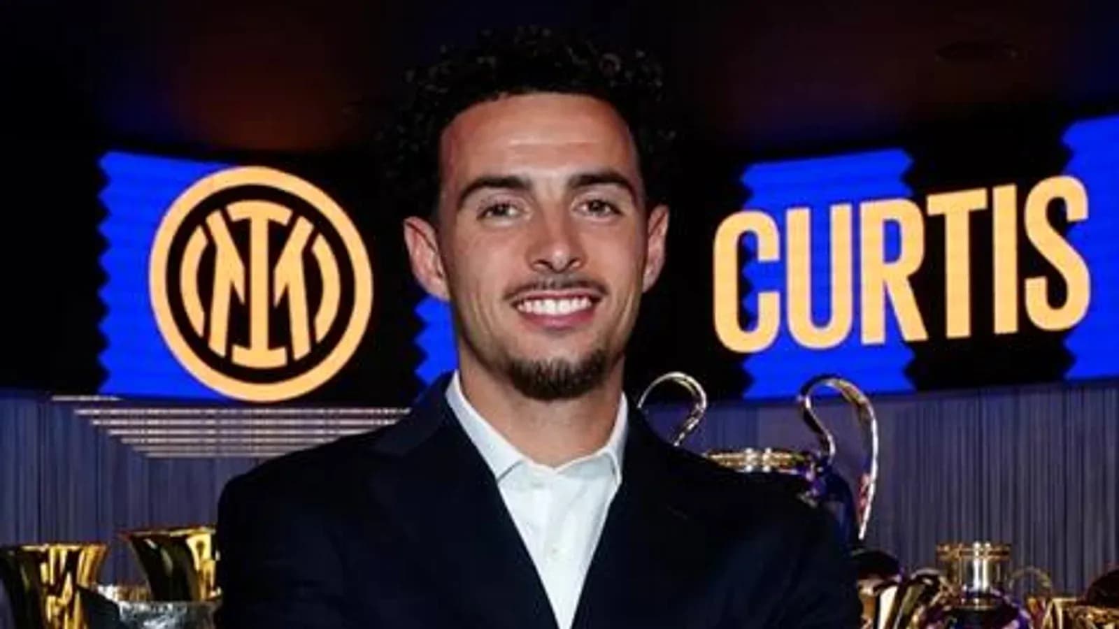 Jugador sonriendo frente a trofeos y logo de Inter Jugador sonriendo frente a trofeos y logo de Inter